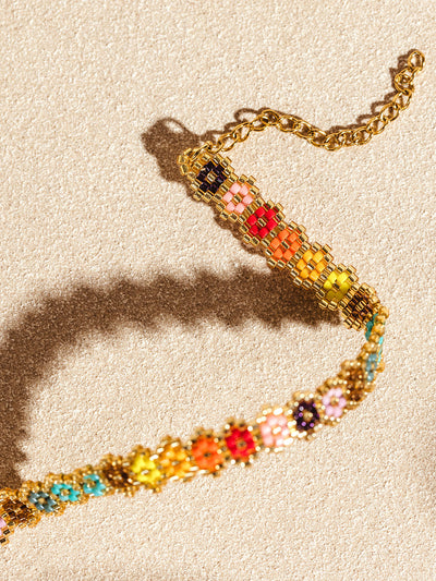 Detailed view of a handcrafted bracelet with colorful beaded flowers, arranged in a gradient pattern, against a sandy beige backdrop. The bracelet's shimmering gold accents and fine beadwork create a striking visual contrast.