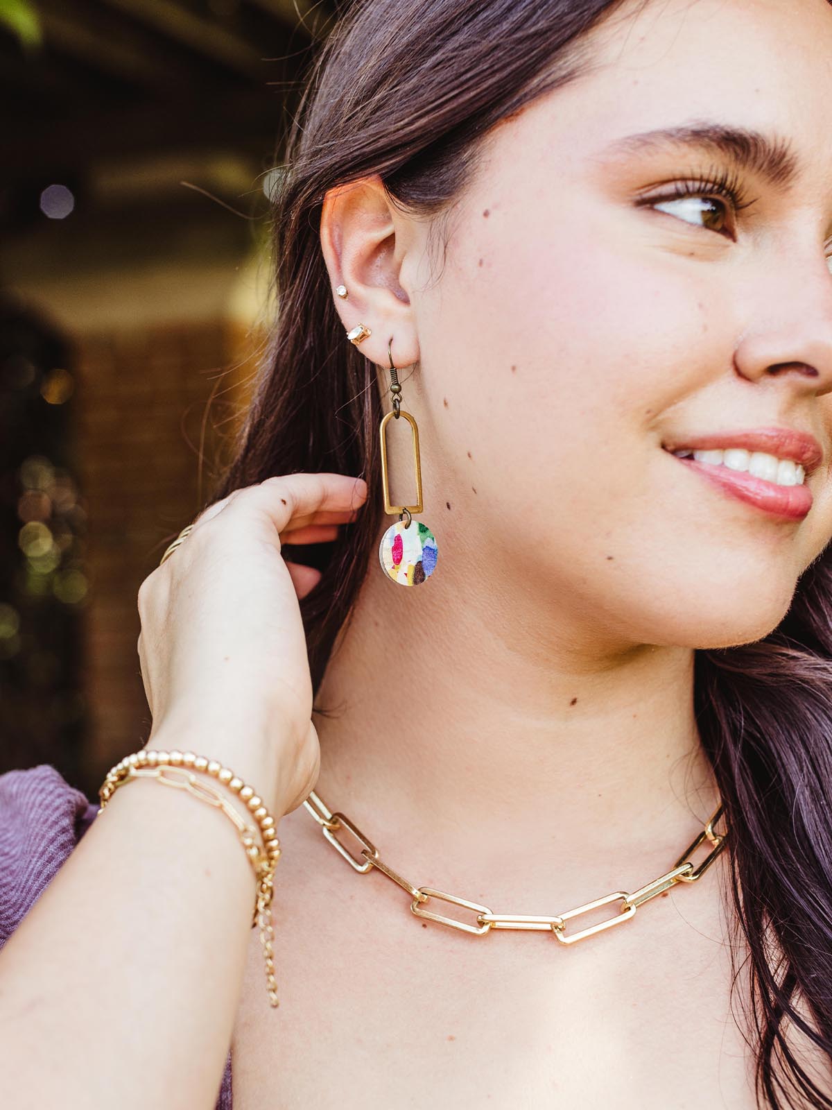 Woman smiling while wearing handcrafted geometric earrings with a brass rectangular frame and a colorful circular pendant, paired with a gold chain necklace.