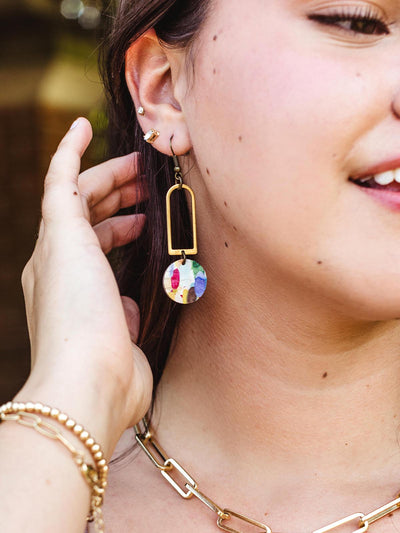 Close-up of a woman adjusting her handcrafted geometric earrings, featuring a brass rectangular frame and a colorful circular pendant, paired with gold jewelry.