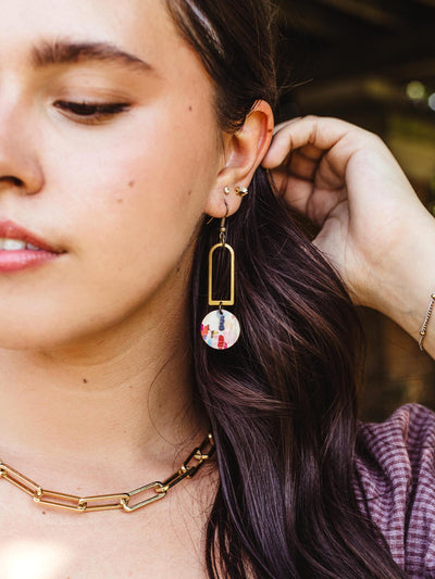 Side profile of a woman wearing handcrafted geometric earrings with a brass rectangular frame and a colorful circular pendant, paired with layered gold jewelry.