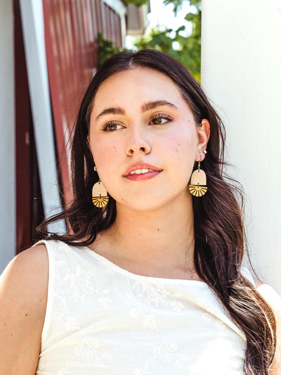 Woman wearing handcrafted white leather and brass fan-shaped earrings, styled with a white embroidered top, in an outdoor setting.