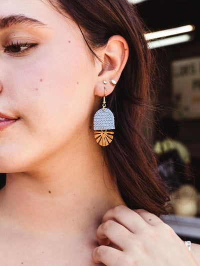 Close-up of a woman wearing handcrafted blue hexagon-pattern leather and brass fan-shaped earrings, styled with other delicate earrings.