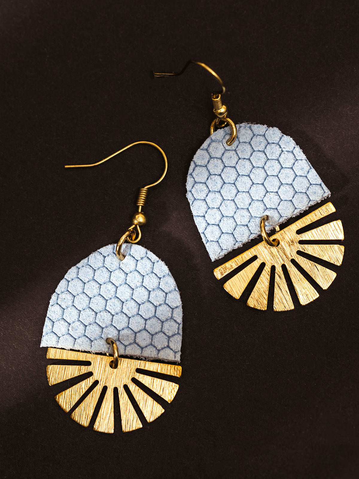 Close-up of handcrafted earrings with blue hexagon-pattern leather and brass fan-shaped accents, displayed on a dark background.