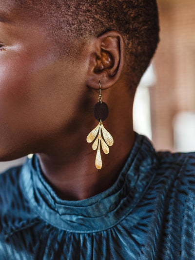 Close-up of a model wearing handcrafted gold leaf-shaped earrings with a black accent, highlighting the detailed design against her blue dress.