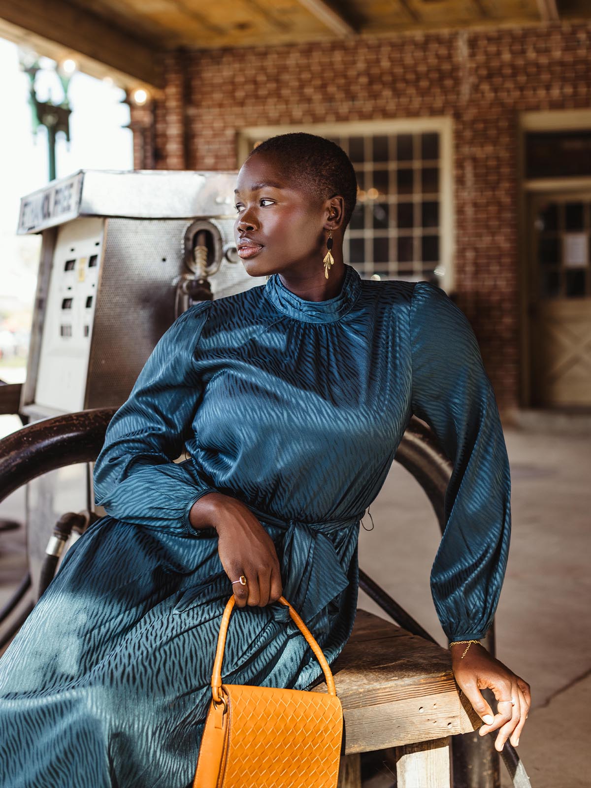 Elegant woman wearing a flowing blue dress, gold leaf-shaped earrings, and holding a woven leather handbag, sitting in a vintage setting with a thoughtful expression.