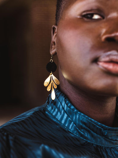 Profile close-up of a model showcasing handcrafted gold leaf-shaped earrings with a black accent, paired with a textured blue dress.