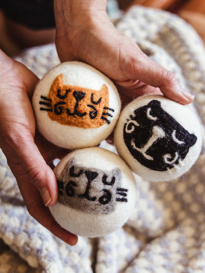 Close-up of hands holding three white wool dryer balls, each hand-felted with a whimsical cat face in black, gray, and orange on a cozy cream and blue knit blanket.