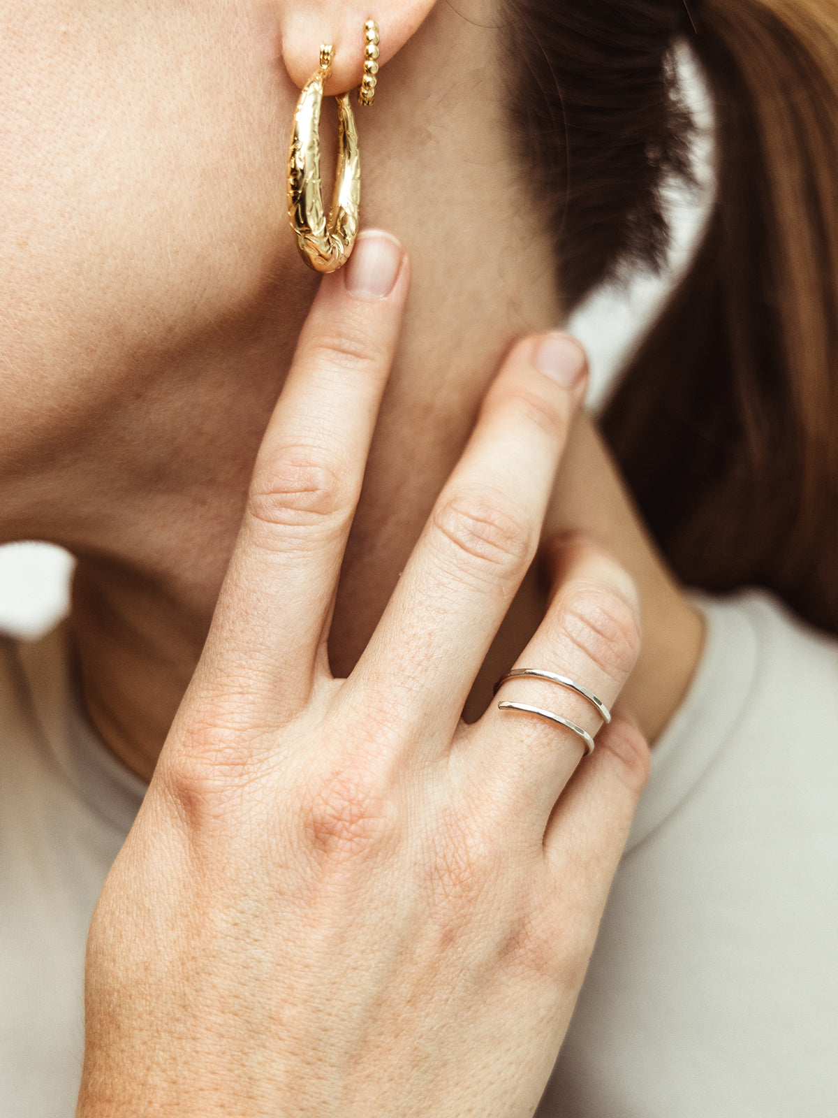 First Image: "Close-up of a woman's hand gently touching her ear, featuring a textured gold hoop earring and a minimalistic silver double band ring on her finger. Perfect for showcasing ethical and handcrafted jewelry for everyday elegance.