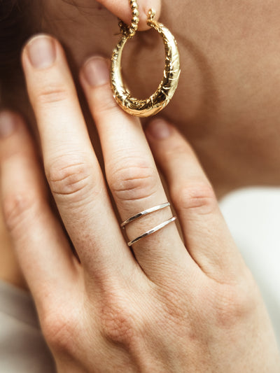 Side view of a woman wearing a textured gold hoop earring paired with a smaller dotted gold hoop earring, her hand resting near her ear, highlighting a sleek silver double band ring. An ideal image for promoting sustainable and artisan-crafted accessories.