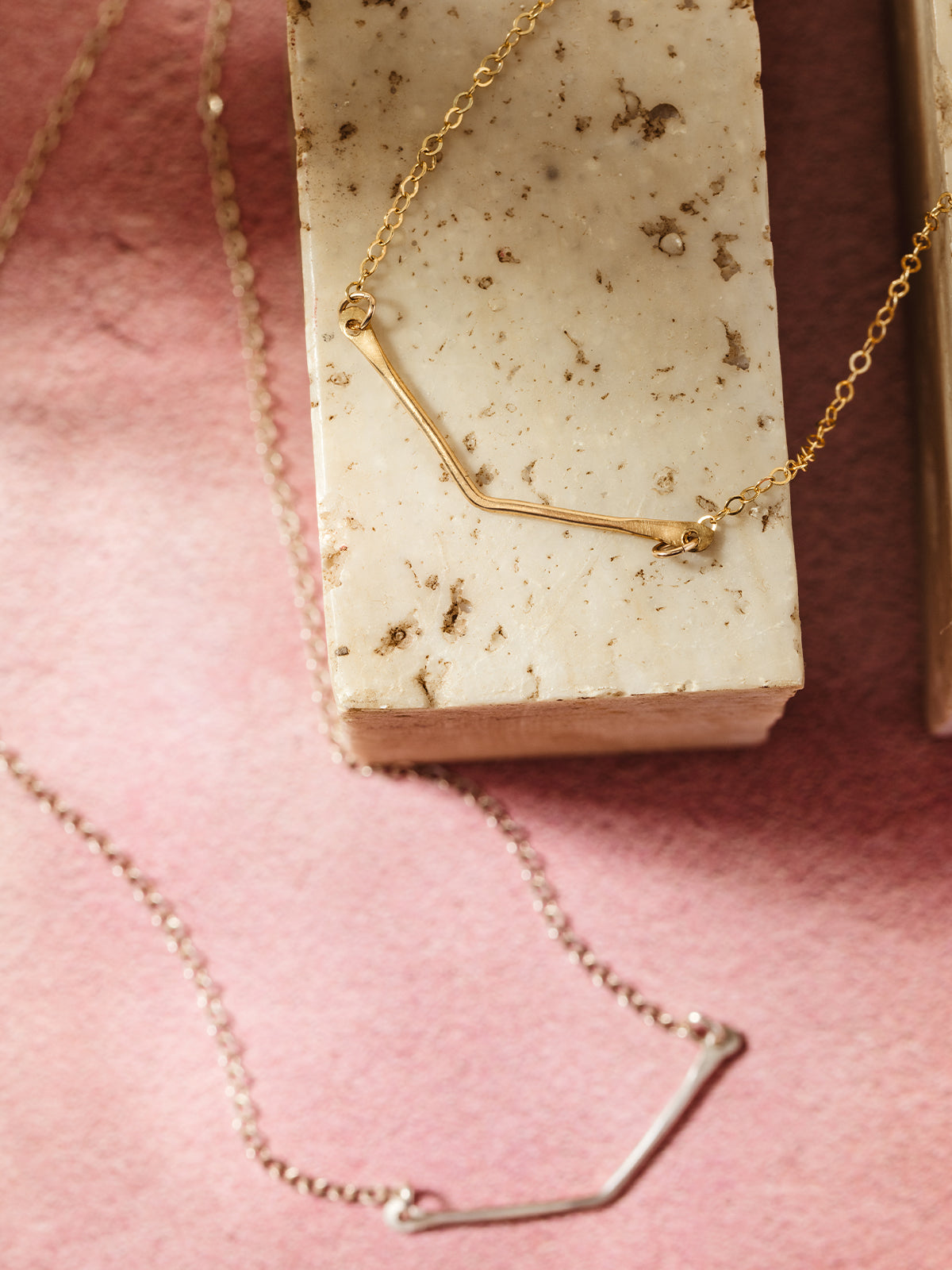 Gold and silver minimalist bar necklaces displayed on natural stone blocks and a blush pink textured background, showcasing elegant handcrafted jewelry with a modern and versatile design.