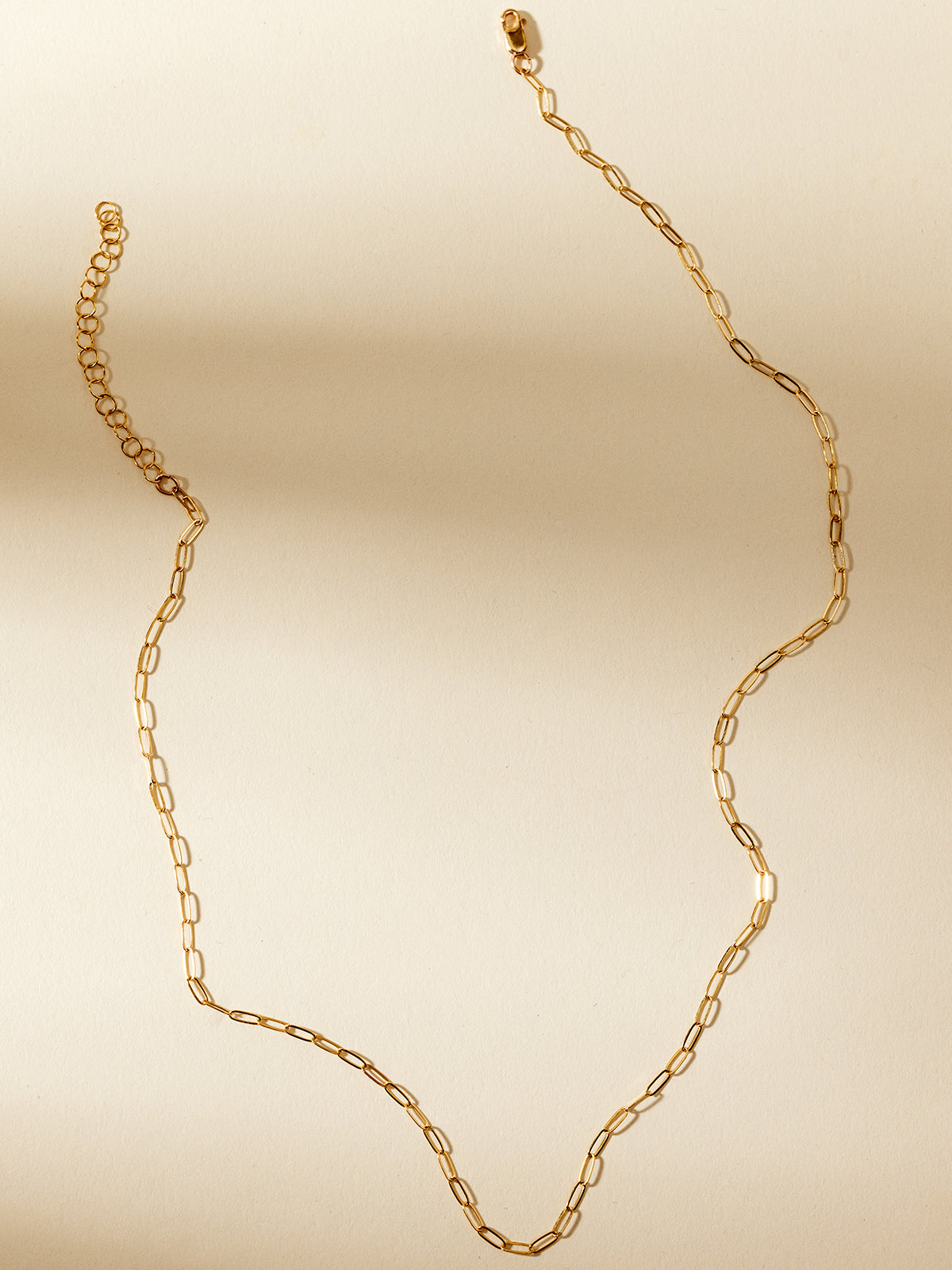 Delicate gold chain necklace with elongated oval links, elegantly displayed on a neutral beige background with soft natural shadows, highlighting minimalist and versatile handcrafted jewelry.