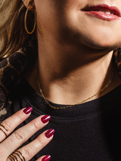 A woman wearing a delicate gold paperclip chain necklace and matching hoop earrings, paired with a black top and a leopard print coat, showcasing a modern and sophisticated jewelry style.