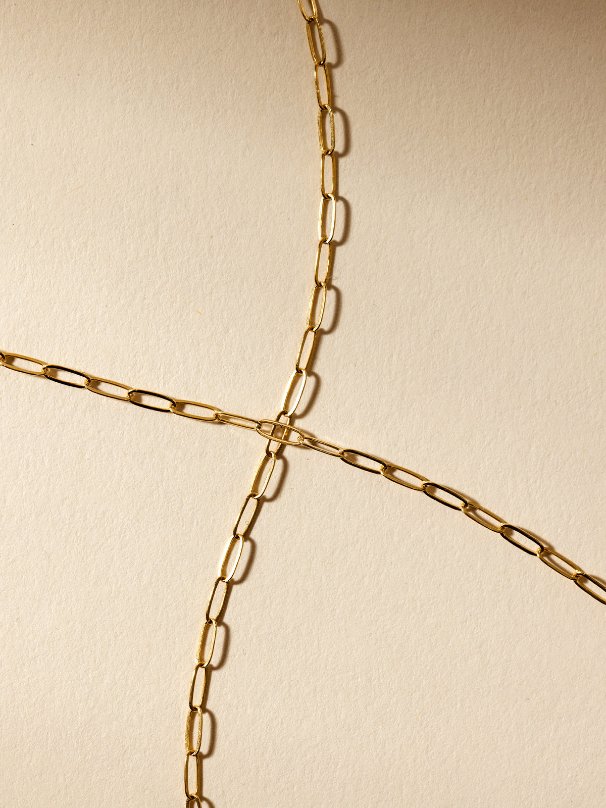 A close-up of a gold paperclip chain necklace displayed on a neutral beige background, highlighting its minimalist design and versatile elegance.