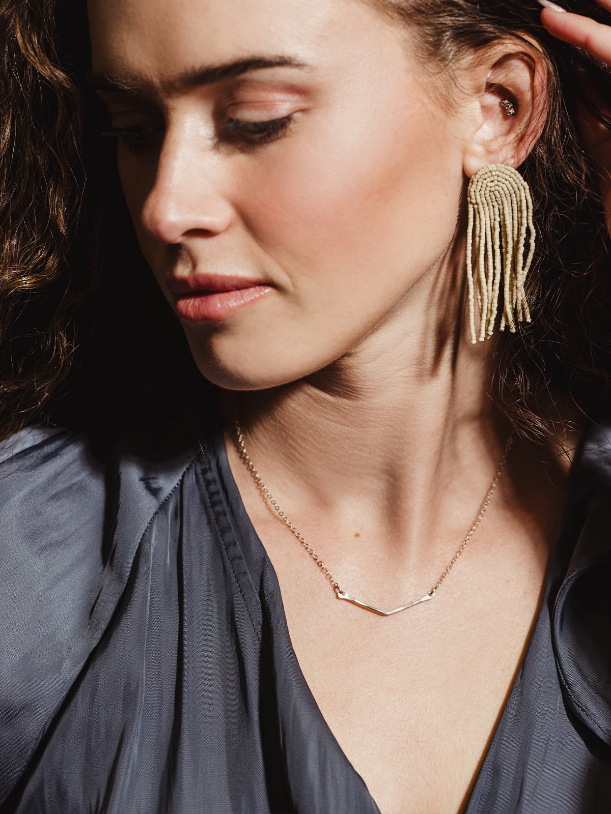 Woman wearing a silver chevron necklace and beaded fringe earrings, looking downward in a soft-lit portrait. The jewelry pieces add an effortless yet sophisticated style to the look.