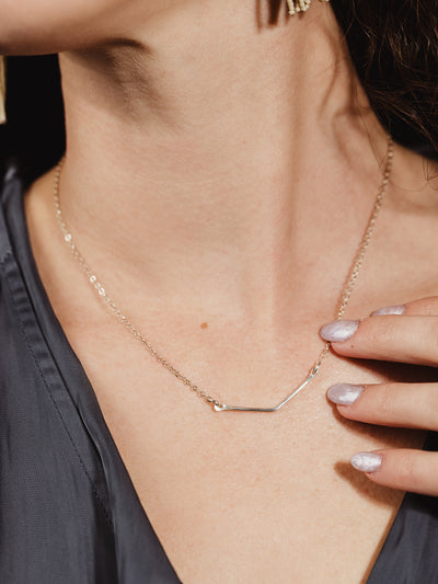 Close-up of a silver chevron necklace on a woman's neck, highlighting the delicate minimalist design. The jewelry is paired with a dark satin blouse, emphasizing the shine and elegance of the piece.