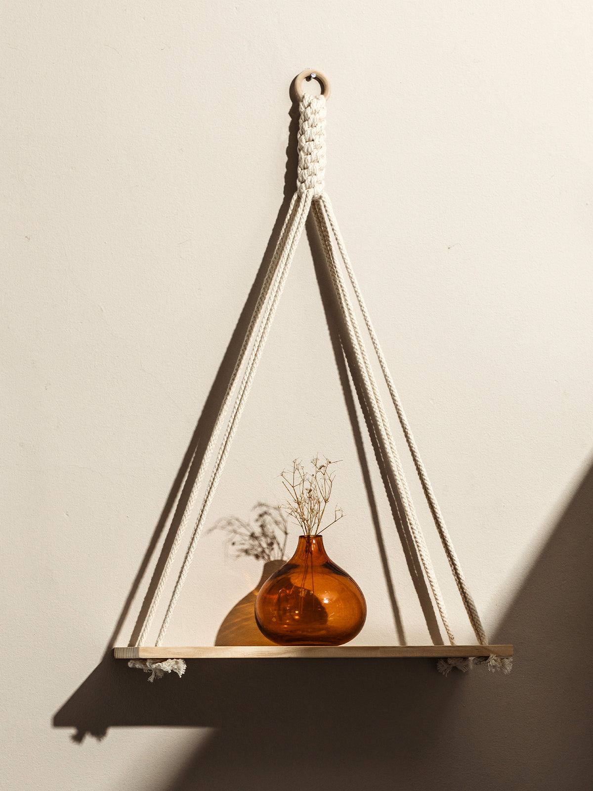 Minimalist wall shelf hanging from braided white rope, holding a small amber glass vase with dried flowers. The shelf casts a soft shadow on the neutral-colored wall, creating a warm and inviting atmosphere.