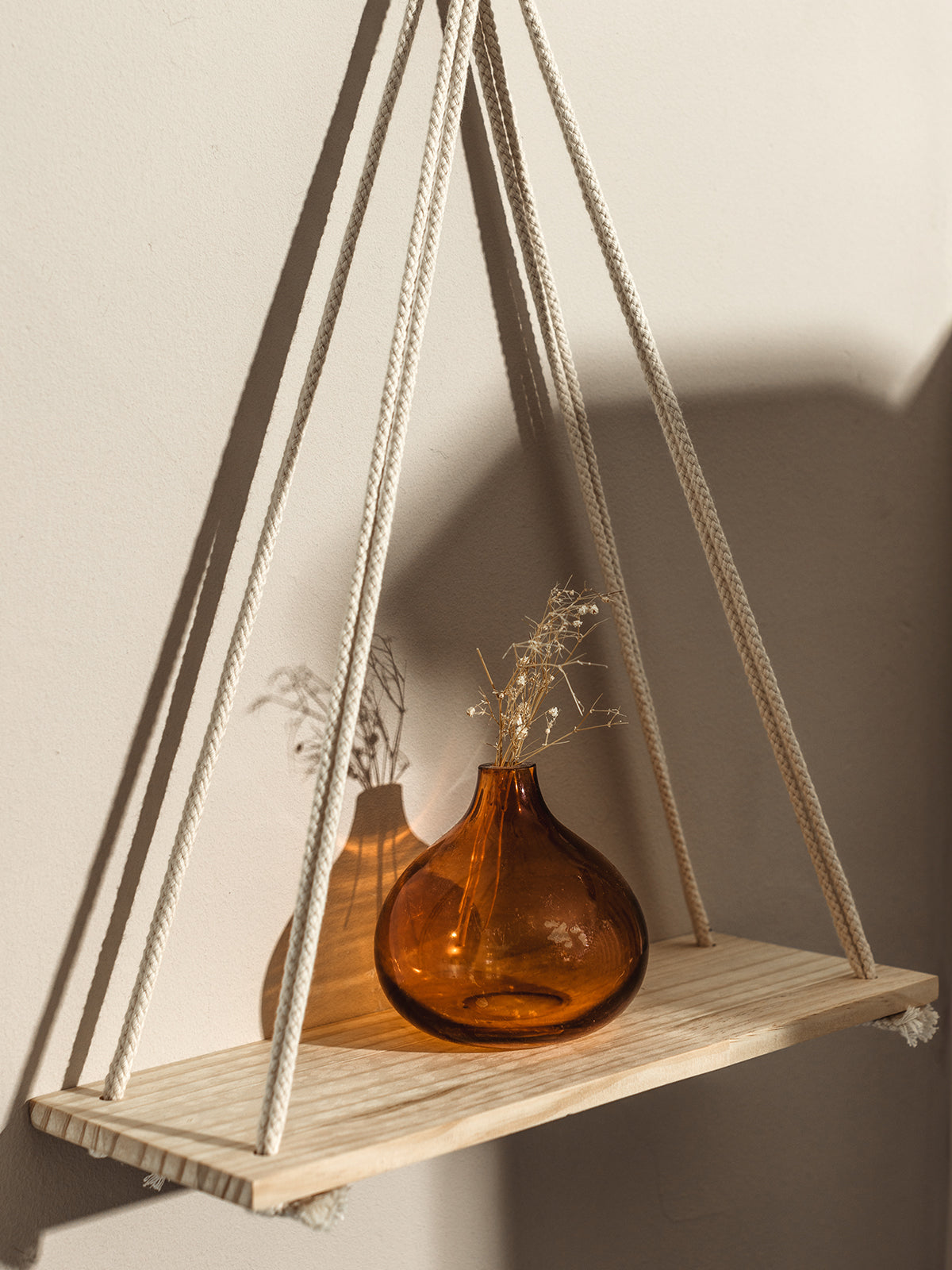 Side view of a simple wall shelf suspended by braided white rope, featuring an amber glass vase with dried flowers. The sunlight streaming in casts delicate shadows, enhancing the natural and earthy aesthetic of the setup.