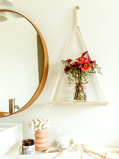 Bright and airy bathroom scene with a wooden-framed round mirror, a hanging shelf displaying a vase with vibrant red and pink flowers, and a stylish arrangement of accessories on the counter, including a peach-colored vase, a candle, and a glass container with cotton pads.