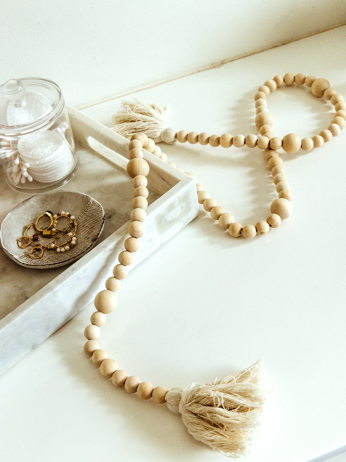 Close-up of a minimalist decor setup featuring a strand of wooden beads with tassels, elegantly laid on a white surface. Nearby, a marble tray holds a small dish with gold rings and a glass jar with cotton pads and swabs.