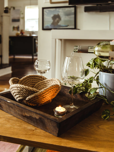 Cozy indoor scene featuring a rustic wooden tray on a table, holding two wine glasses, one being filled with white wine. A small lit candle and a decorative woven basket add warmth, while a leafy houseplant in the background enhances the inviting atmosphere.