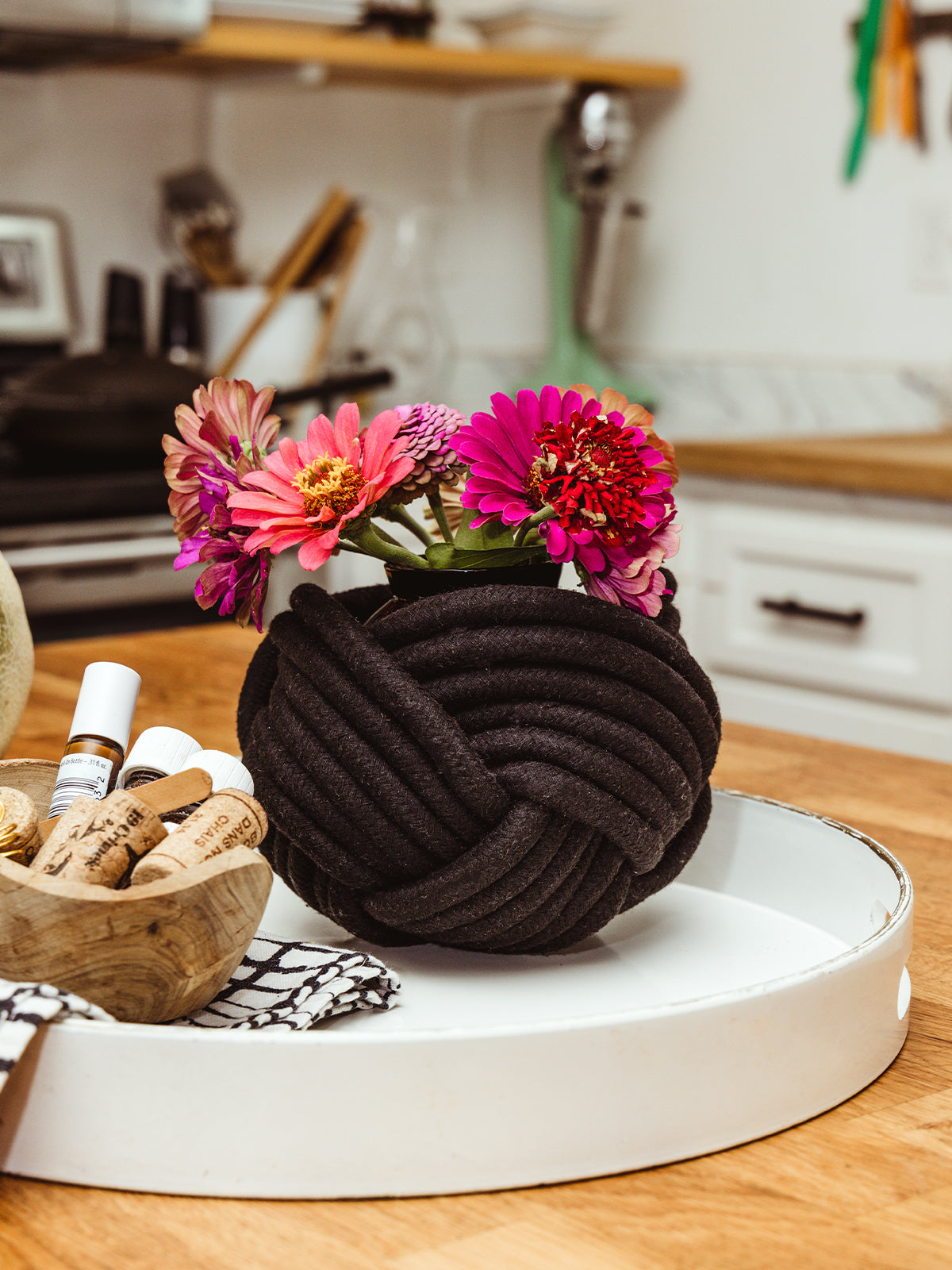 Bright kitchen scene featuring a small bouquet of colorful zinnia flowers in a black, braided fabric-wrapped vase, placed on a white tray. Nearby, a wooden bowl holds corks and essential oil bottles, adding a touch of rustic charm to the modern kitchen countertop.