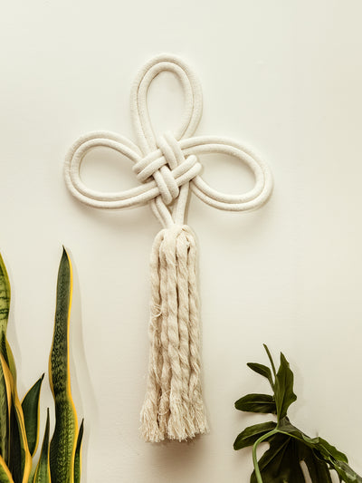 Wall-mounted decorative piece made of white braided rope intricately tied into a knot design with loops and a tassel hanging down, set against a neutral background with green snake plants and other foliage framing the bottom of the image.