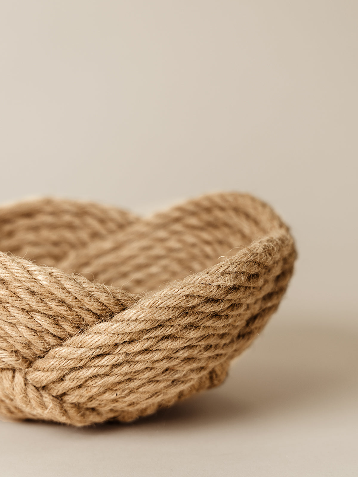 Close-up of a handwoven basket made from natural rope, showcasing a complex, braided pattern. The basket is set against a neutral beige background, highlighting its earthy texture and craftsmanship.