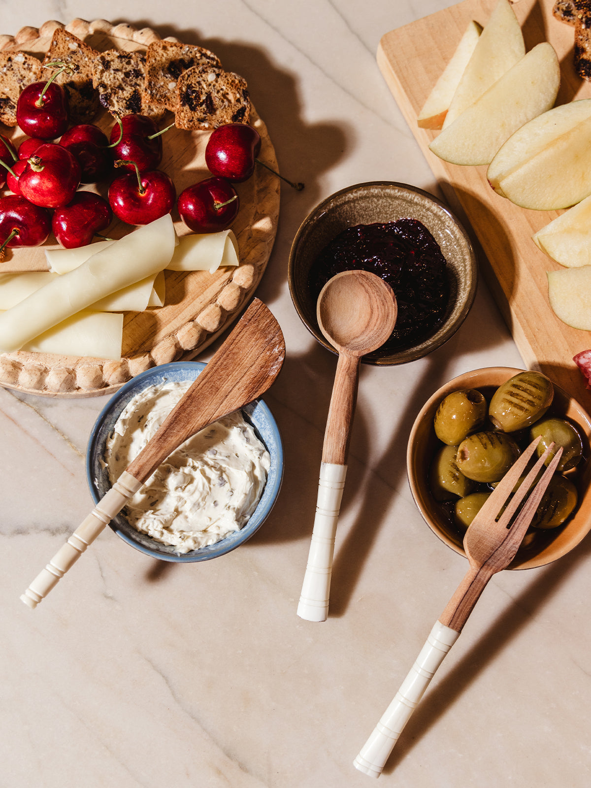 Appetizer serving utensils on white counter with charcuterie foods and spreads.