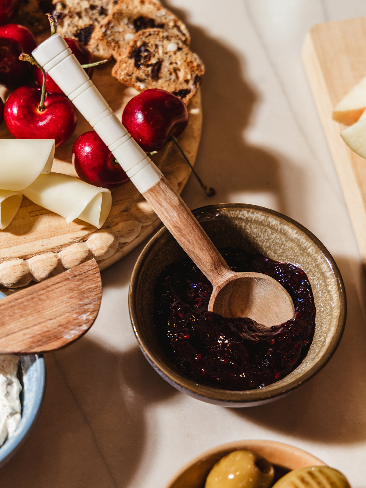 Appetizer serving spoon on white counter with charcuterie foods and spreads.