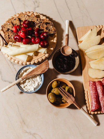 Appetizer serving utensils on white counter with charcuterie foods and spreads.