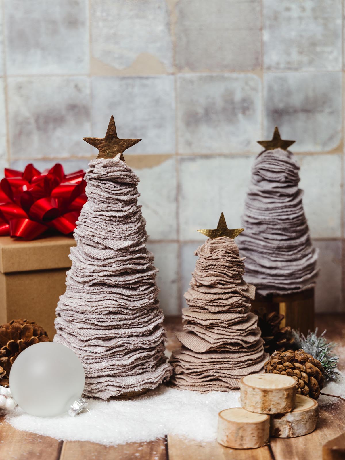 Set of three handcrafted fabric Christmas trees with brass star toppers in different sizes, surrounded by festive decor including pinecones, a red bow, and wood slices. Rustic holiday decorations for creating a cozy and handmade holiday atmosphere.