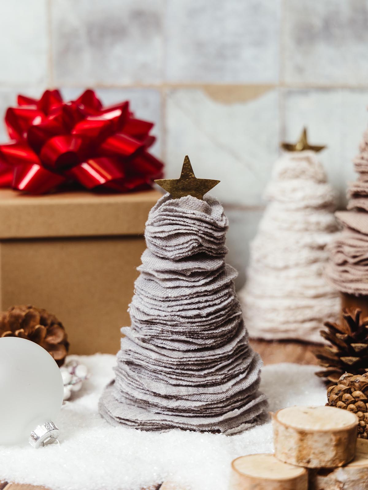 Small handcrafted gray fabric Christmas tree with a brass star topper, surrounded by festive decorations including a red bow and pinecones. Perfect rustic holiday decor for a cozy home atmosphere.