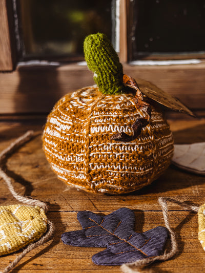 Crocheted pumpkin on wood table near window with fall leaf garland layer around it. 