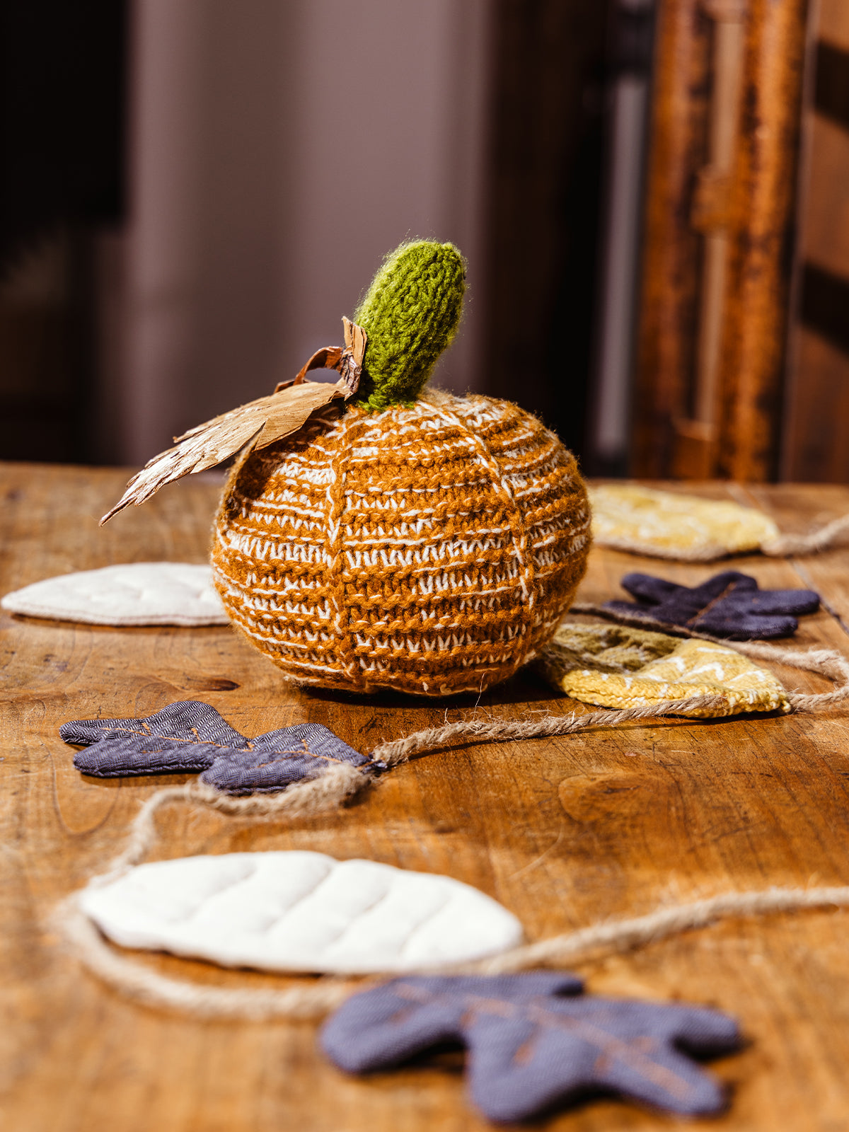 Crocheted pumpkin on wood table near window with fall leaf garland layer around it. 