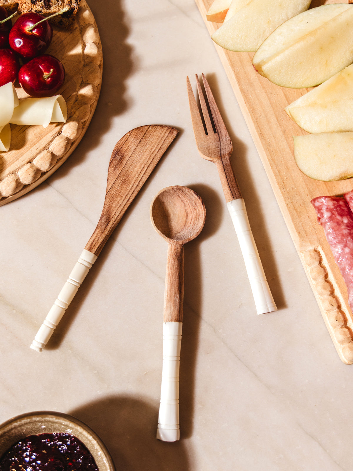 Appetizer serving utensils on white counter with charcuterie foods and spreads.