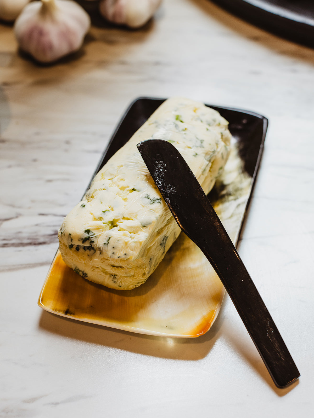 Butter tray and knife with garlic butter atop tray. Placed on a white kitchen counter with whole garlic. 