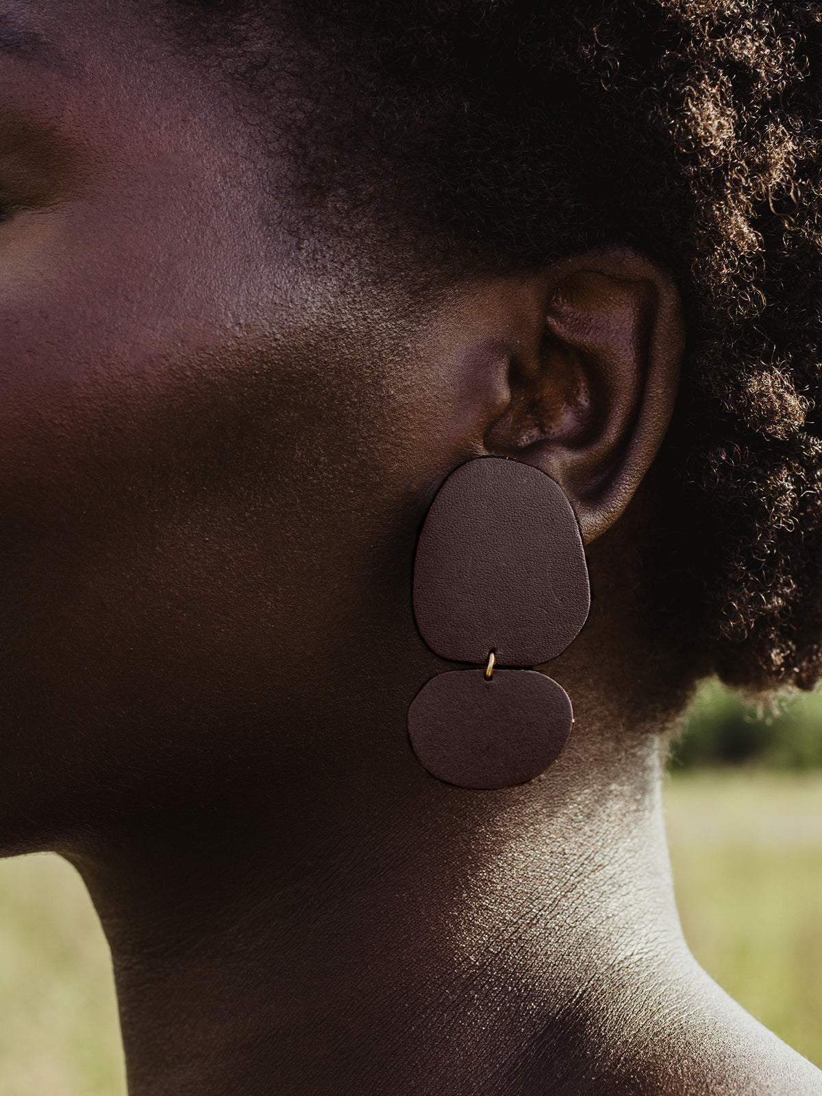 Close-up of a woman wearing brown leather drop earrings, emphasizing the texture of the handcrafted design against a natural outdoor background.