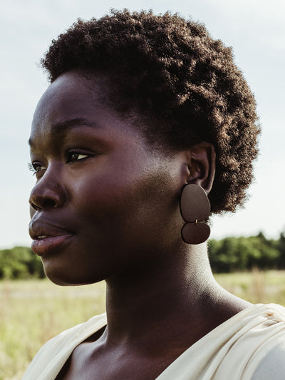 Side profile of a woman with short curly hair wearing handcrafted brown leather drop earrings, photographed outdoors in a natural setting.