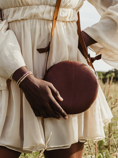 Closeup of female African model in cream dress holding the canteen purse with her hand while the purse is over her shoulder. Model and purse are in an outdoor setting. 