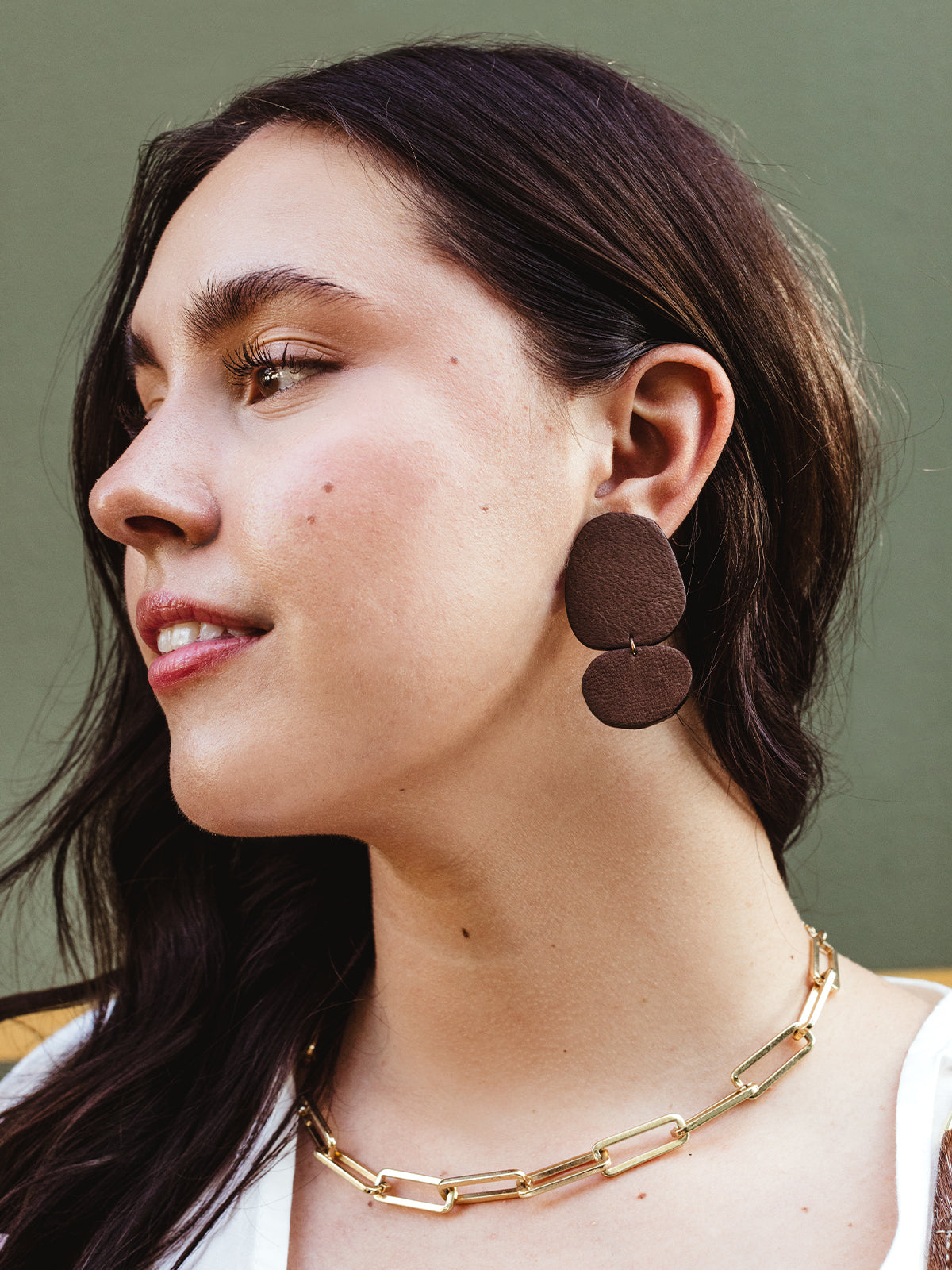 Side profile of a woman with long dark hair wearing handcrafted brown leather drop earrings and a gold chain necklace, set against a green background.