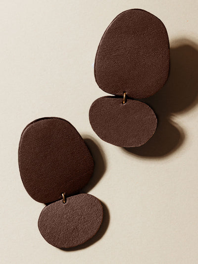 Close-up of a pair of handcrafted brown leather drop earrings with a smooth texture, displayed on a neutral-colored surface.