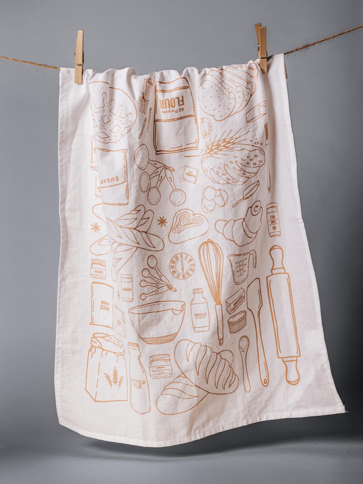 Tea towel with intricate orange line drawings of kitchen and baking essentials, including flour, rolling pin, and various ingredients, hanging on a clothesline with wooden clothespins against a gray background.