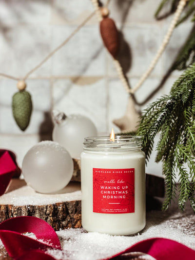 Natural soy candle in a glass jar labeled 'Waking Up Christmas Morning' by Highland Ridge Decor, surrounded by faux snow, greenery, and festive ornaments, creating a cozy holiday atmosphere.