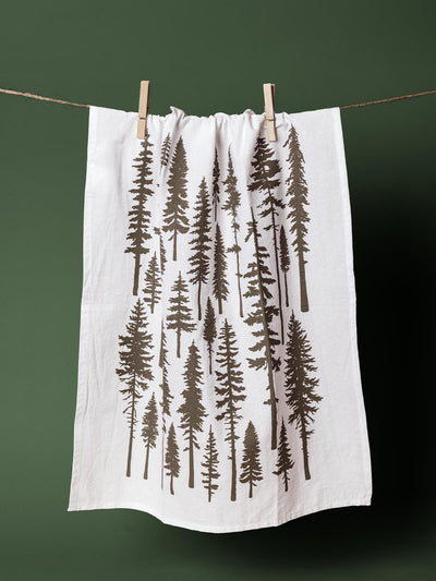 White kitchen towel with a printed pattern of green evergreen trees, hanging on a clothesline with a green background, ideal for rustic and nature-inspired holiday decor.