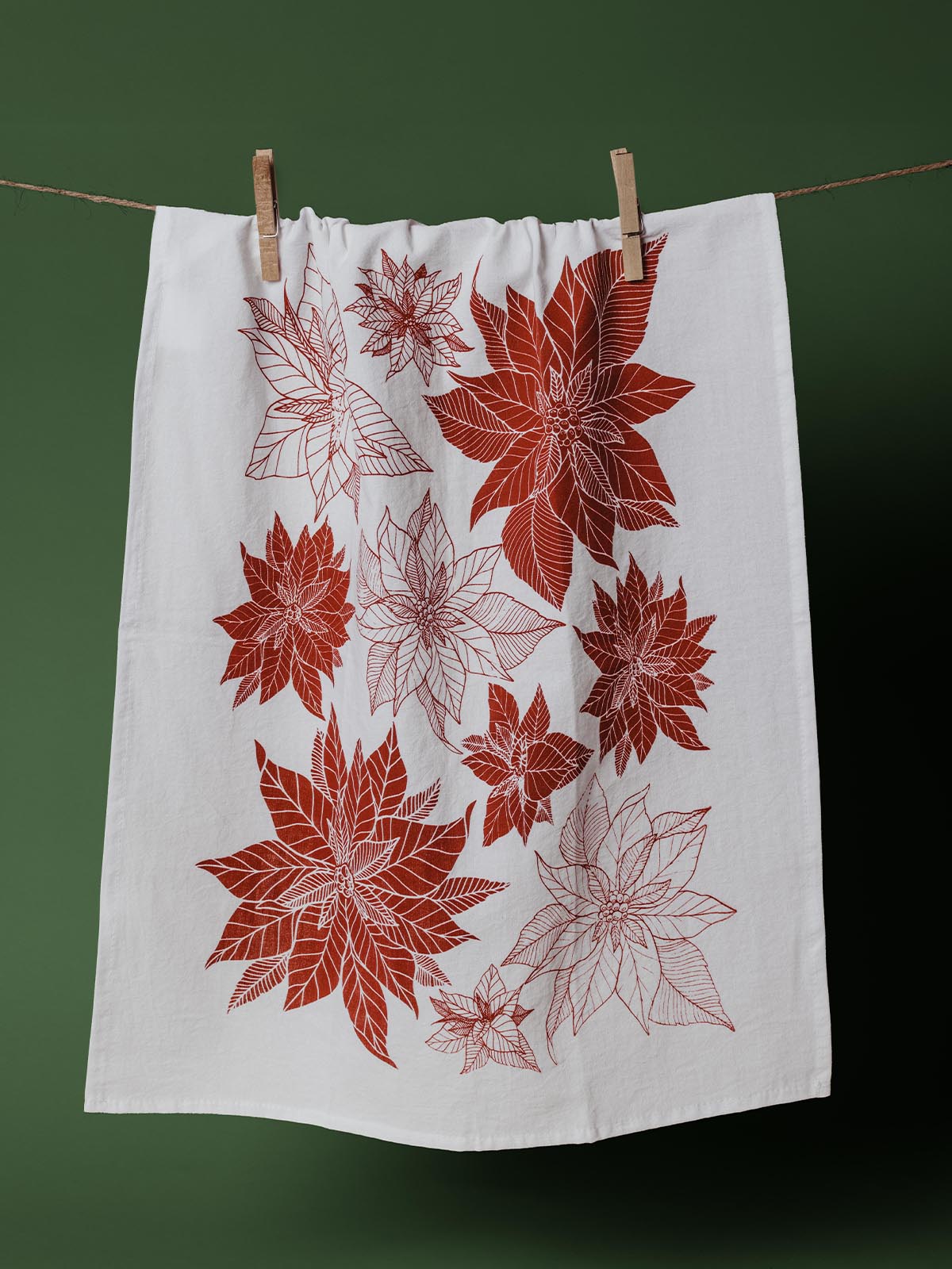 White kitchen towel featuring a red poinsettia print, hanging on a clothesline with a green background, perfect for holiday decor and festive kitchens.