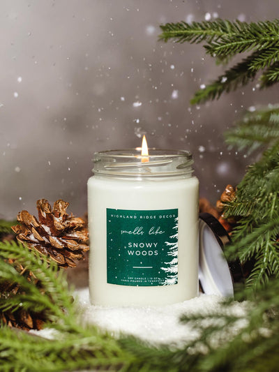 Hand-poured soy candle labeled 'Smells Like Snowy Woods' by Highland Ridge Decor, surrounded by pine branches and pinecones, with snow falling in the background. 