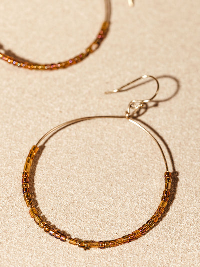 Close-up of handcrafted hoop earrings with intricate brown and gold beads, displayed on a soft, neutral surface.