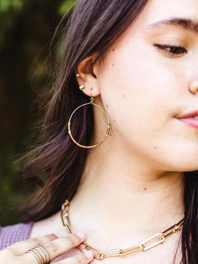 Close-up of a woman wearing handcrafted hoop earrings with brown and gold beads, paired with gold jewelry, against a blurred outdoor background.