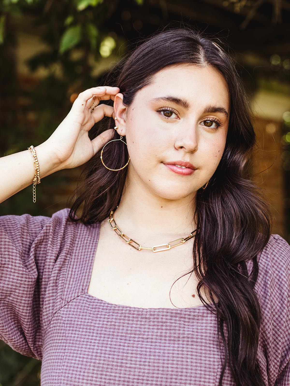 Woman with long dark hair wearing handcrafted hoop earrings with brown and gold beads, a gold chain necklace, and a purple gingham dress, posing outdoors with a hand adjusting her earring.