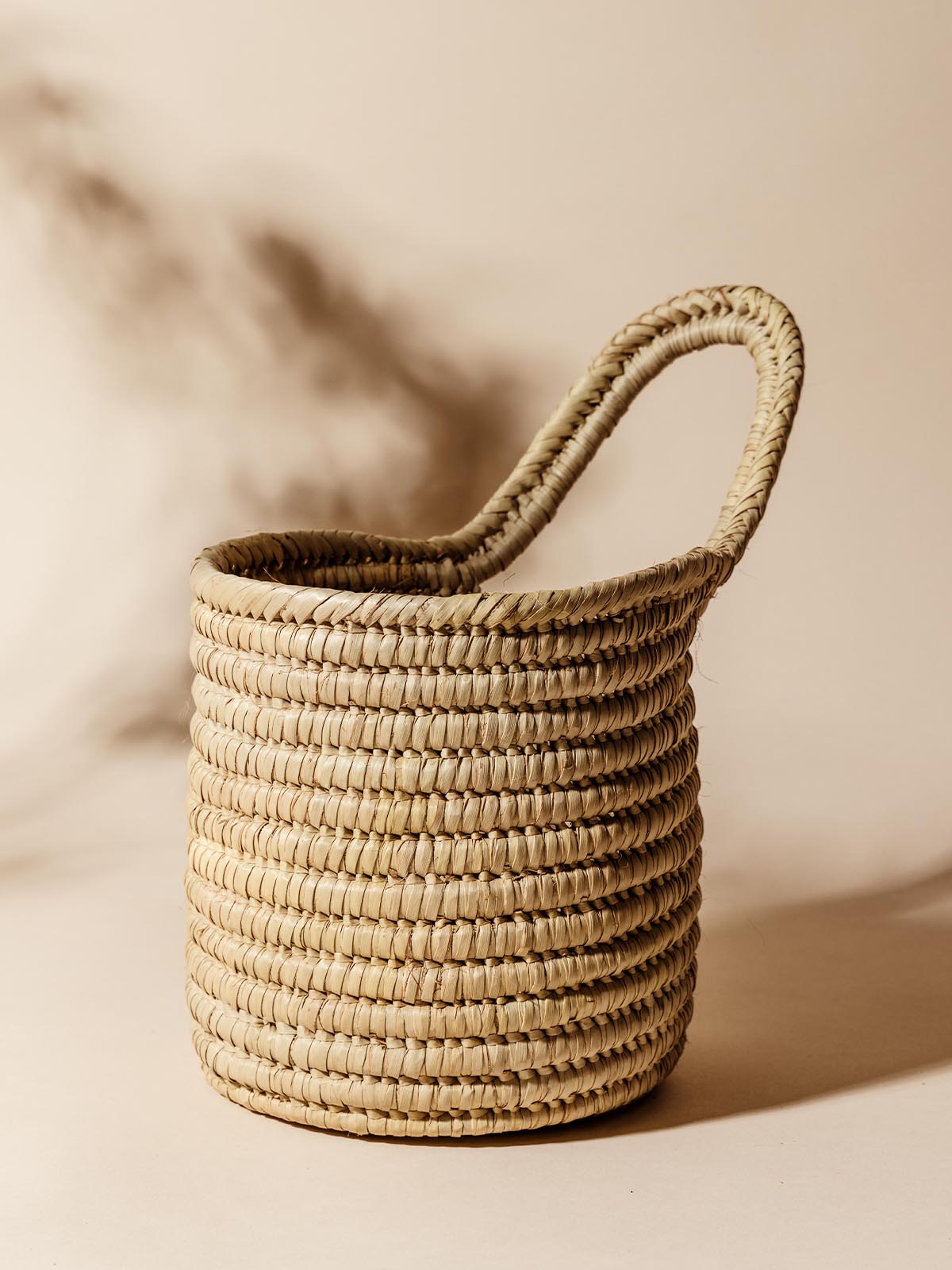 Close-up of a handcrafted woven basket with a single looped handle, placed against a neutral beige background, highlighting its textured natural fibers.